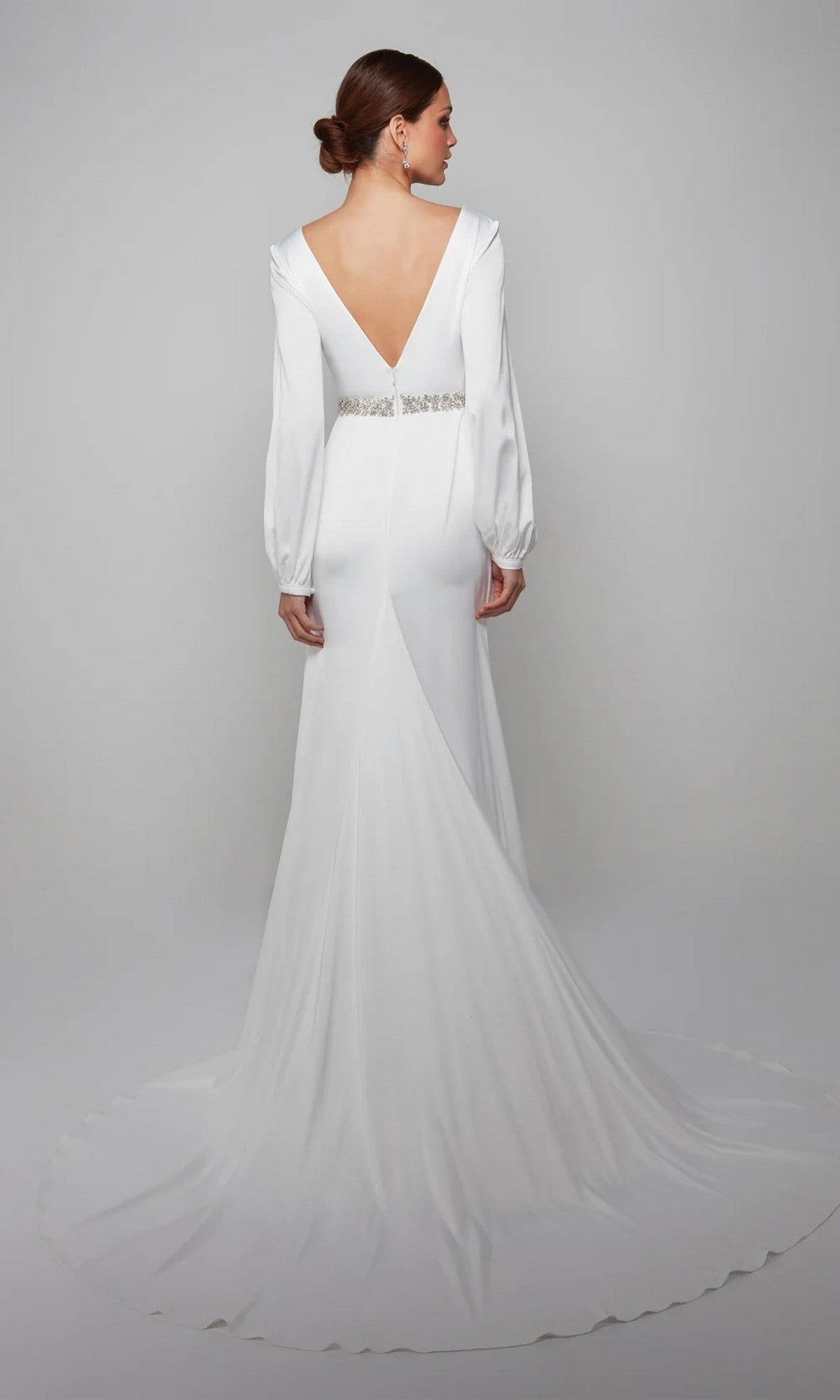 A woman stands facing away against a plain gray background, wearing the Alyce 7070 long formal dress—a white satin gown with long sleeves, an open back, embellished waistband, floor-length silhouette, and slight train.